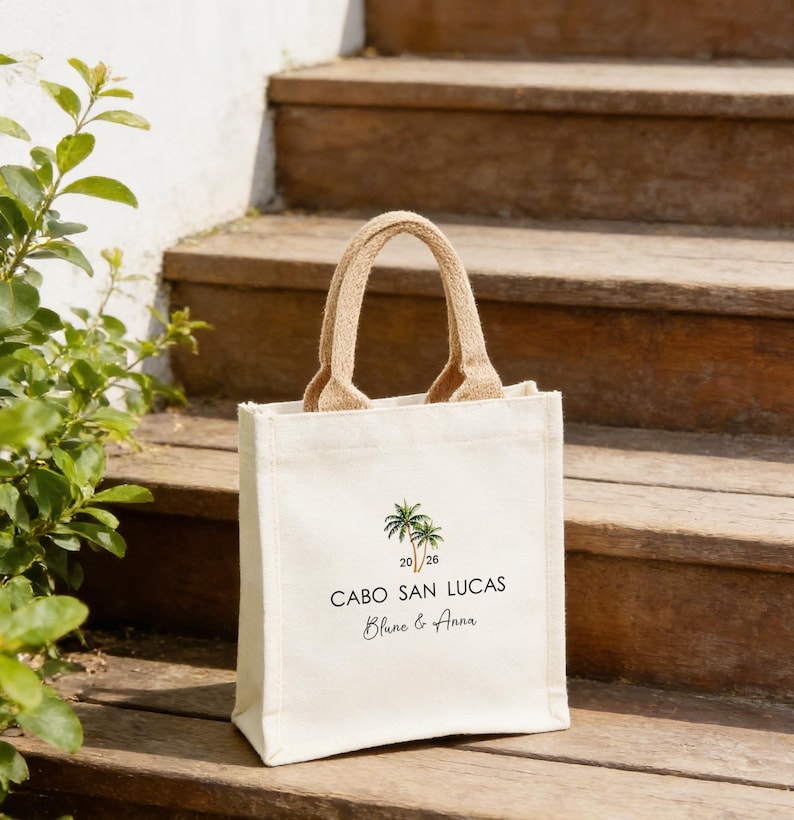 May include: An off-white canvas tote bag with a jute handle. The bag has a palm tree graphic and the text "CABO SAN LUCAS" with the names "Blane & Anna" below. The bag is placed on wooden steps.