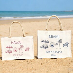 May include: Two off-white canvas tote bags with beige handles sit on a sandy beach. One bag says "HAWAII" in maroon with beach-themed illustrations. The other says "MIAMI" in navy blue with similar illustrations.