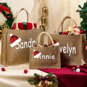 May include: Three burlap tote bags with white text and Santa hats. The bags are personalized with the names Sandra, Annie, and Velyn. The bags are filled with gifts and are set against a Christmas background.