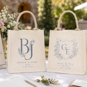 May include: Two off-white canvas tote bags with rounded handles. The left bag features the blue monogram BJ, the text BONNIE & JOHN, and 9.20.2026. The right bag features the blue monogram GE inside a floral crest, the text Gary & Eva, and 10.2.2026.