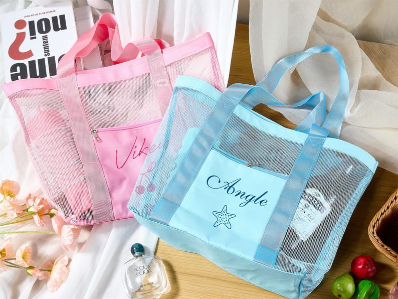 May include: Two mesh tote bags, one pink and one blue, with matching handles and front pockets. The pink bag has the name "Vikey" and a cherry design, while the blue bag says "Angle" with a starfish. Both bags are filled with items.
