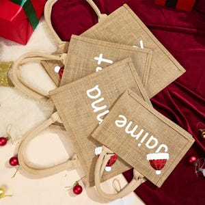 May include: Tan burlap tote bags with white text and Santa hat graphics. The bags have beige handles and are arranged on a red velvet surface. Christmas-themed gift bags.