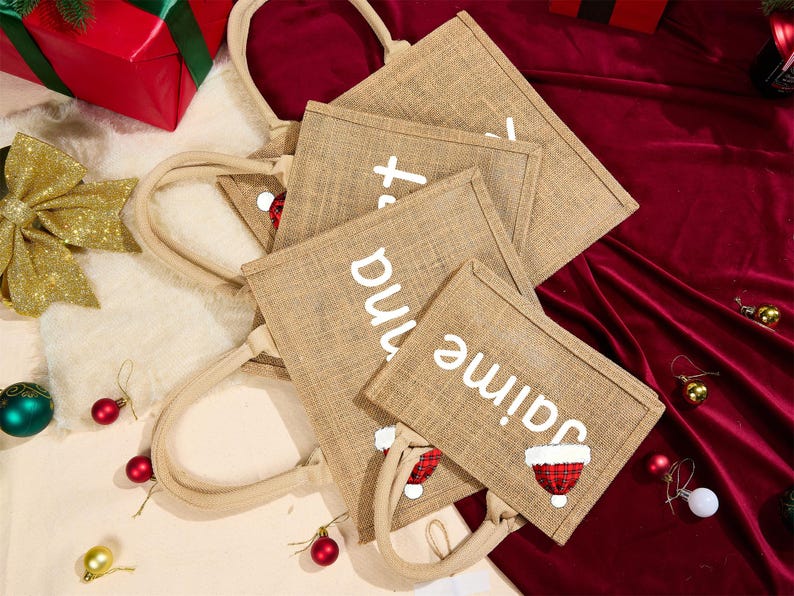 May include: Tan burlap tote bags with white text and Santa hat graphics. The bags have beige handles and are arranged on a red velvet surface. Christmas-themed gift bags.