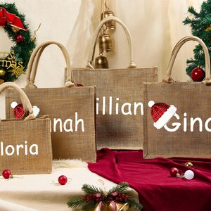 May include: Four burlap tote bags with white text and Santa hats. The bags are personalized with the names "Gloria", "Hannah", "Lillian", and "Gina". A Christmas wreath and tree with ornaments are in the background.