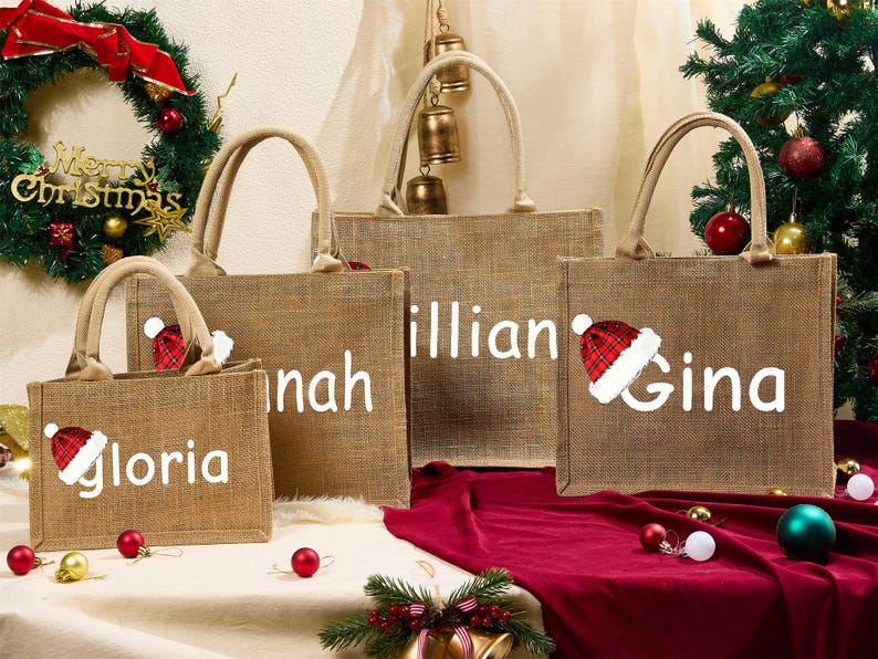 May include: Four burlap tote bags with white text and Santa hats. The bags are personalized with the names "Gloria", "Hannah", "Lillian", and "Gina". A Christmas wreath and tree with ornaments are in the background.