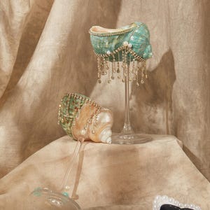 May include: Two decorative wine glasses with seashell bowls. One glass stands upright, the other is tilted. The shells are teal and cream, adorned with pearls and dangling crystal accents. The glasses sit on a beige surface against a neutral backdrop.
