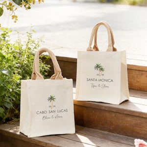 May include: Two cream-coloured canvas tote bags with jute handles. Each bag features a palm tree graphic and text. One says "Cabo San Lucas" and the other says "Santa Monica". The bags are on wooden steps.