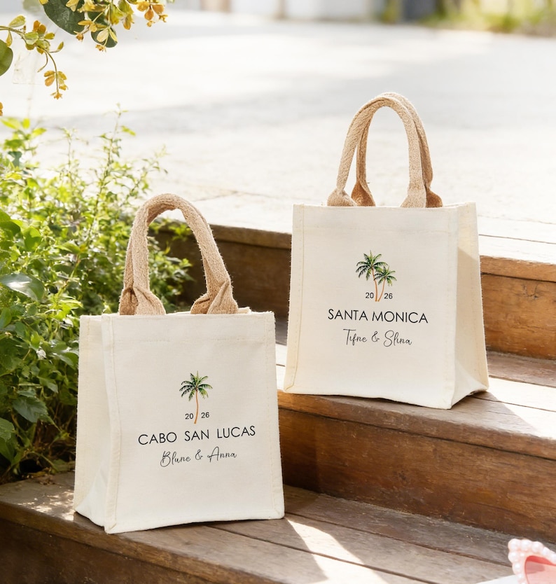 May include: Two cream-coloured canvas tote bags with jute handles. Each bag features a palm tree graphic and text. One says "Cabo San Lucas" and the other says "Santa Monica". The bags are on wooden steps.