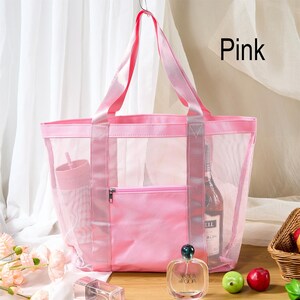 May include: Pink mesh tote bag with a solid pink front pocket and handles. The bag is hanging from a silver hook. The word "Pink" is written in black text in the upper right corner. A bottle of perfume and some fruit are in front of the bag.