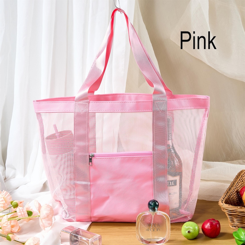 May include: Pink mesh tote bag with a solid pink front pocket and handles. The bag is hanging from a silver hook. The word "Pink" is written in black text in the upper right corner. A bottle of perfume and some fruit are in front of the bag.