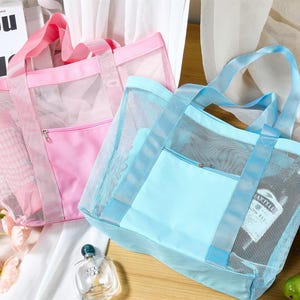 May include: Two mesh tote bags, one pink and one light blue, with matching handles and zippered front pockets. The bags are partially transparent, revealing the contents inside. The bags are on a white surface.