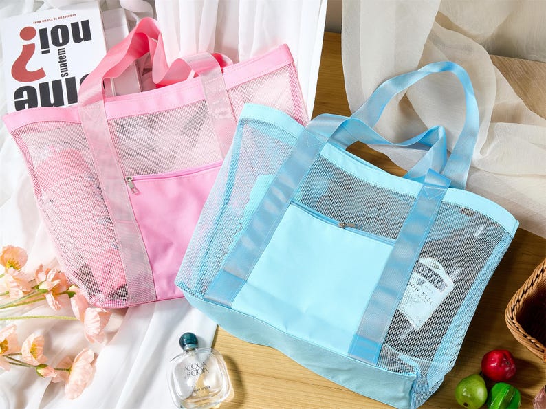May include: Two mesh tote bags, one pink and one light blue, with matching handles and zippered front pockets. The bags are partially transparent, revealing the contents inside. The bags are on a white surface.