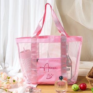 May include: A pink mesh tote bag with matching handles and a front pocket. The pocket features the name "Annea" and a watermelon slice graphic. The bag contains a tumbler, a bottle of wine, and other items. The bag is designed for beach or travel.