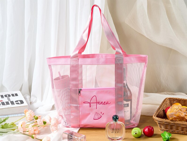 May include: A pink mesh tote bag with matching handles and a front pocket. The pocket features the name "Annea" and a watermelon slice graphic. The bag contains a tumbler, a bottle of wine, and other items. The bag is designed for beach or travel.