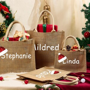May include: Several burlap tote bags with Christmas-themed designs. Each bag has a name printed on it: Stephanie, Mildred, Linda, and Paula. A Christmas wreath and tree are in the background, with gifts and decorations.