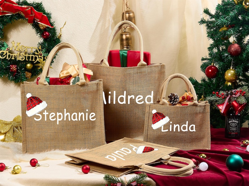 May include: Several burlap tote bags with Christmas-themed designs. Each bag has a name printed on it: Stephanie, Mildred, Linda, and Paula. A Christmas wreath and tree are in the background, with gifts and decorations.