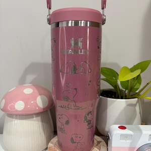 May include: A pink Stanley tumbler with a handle and lid, featuring Snoopy character illustrations. The tumbler has the Stanley logo and is surrounded by a ceramic mushroom, a small camera, and a plant in a white pot.