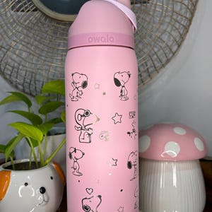 Puede incluir: Una botella de agua rosa Owala con ilustraciones de Snoopy y el nombre de la marca. La botella tiene una tapa rosa y una correa. El dise&ntilde;o incluye ilustraciones de Snoopy, estrellas y corazones. Capacidad aproximada de 700 ml.