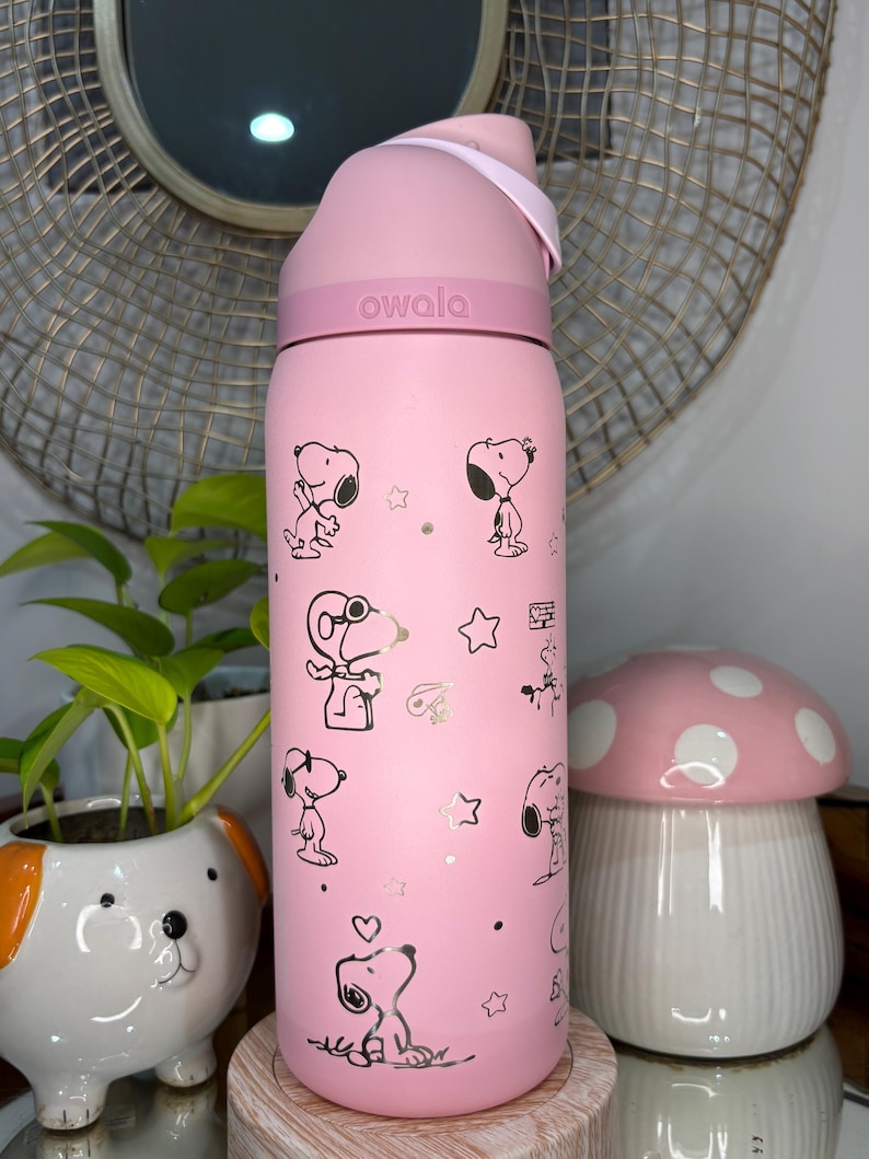Puede incluir: Una botella de agua rosa Owala con ilustraciones de Snoopy y el nombre de la marca. La botella tiene una tapa rosa y una correa. El dise&ntilde;o incluye ilustraciones de Snoopy, estrellas y corazones. Capacidad aproximada de 700 ml.