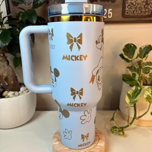 Puede incluir: Un vaso azul claro con dise&ntilde;os dorados de Mickey Mouse y lazos. El vaso tiene un asa y una tapa transparente con borde dorado. La palabra "Mickey" est&aacute; impresa en dorado. El vaso est&aacute; sobre un posavasos de madera.