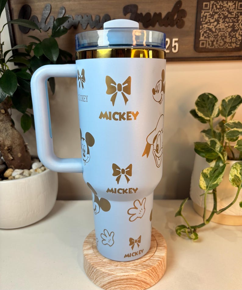 Puede incluir: Un vaso azul claro con dise&ntilde;os dorados de Mickey Mouse y lazos. El vaso tiene un asa y una tapa transparente con borde dorado. La palabra "Mickey" est&aacute; impresa en dorado. El vaso est&aacute; sobre un posavasos de madera.