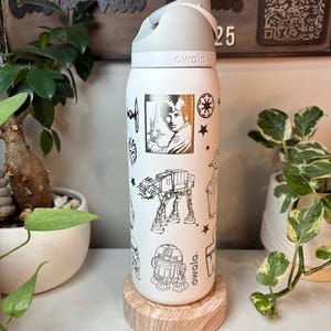 May include: White Owala water bottle with a gray lid and handle, featuring black line art of Star Wars characters and symbols. The bottle is decorated with images of Luke Skywalker, R2-D2, and an AT-AT walker. The bottle is sitting on a wooden coaster.