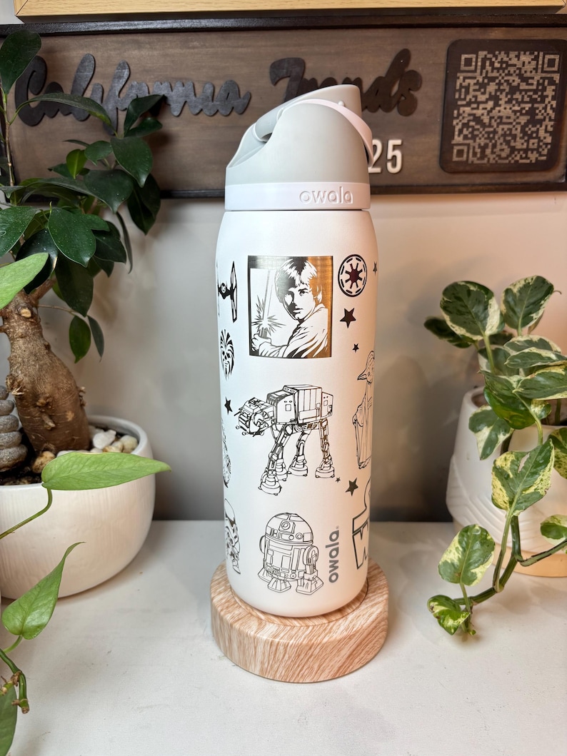 May include: White Owala water bottle with a gray lid and handle, featuring black line art of Star Wars characters and symbols. The bottle is decorated with images of Luke Skywalker, R2-D2, and an AT-AT walker. The bottle is sitting on a wooden coaster.
