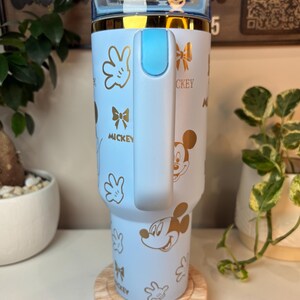 Puede incluir: Un vaso azul claro con dise&ntilde;os dorados de Mickey Mouse y la palabra "Mickey". El vaso tiene un asa y tapa azul claro con borde dorado. Se muestra sobre un posavasos de madera.