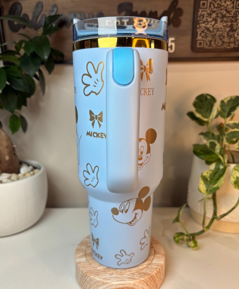 Puede incluir: Un vaso azul claro con dise&ntilde;os dorados de Mickey Mouse y la palabra "Mickey". El vaso tiene un asa y tapa azul claro con borde dorado. Se muestra sobre un posavasos de madera.