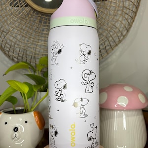 Puede incluir: Botella de agua Owala blanca con ilustraciones de Snoopy en negro y dorado. La botella tiene una tapa rosa y una banda verde claro. El logotipo de Owala est&aacute; impreso en amarillo. Una seta decorativa y una maceta con forma de perro est&aacute;n en el fondo.