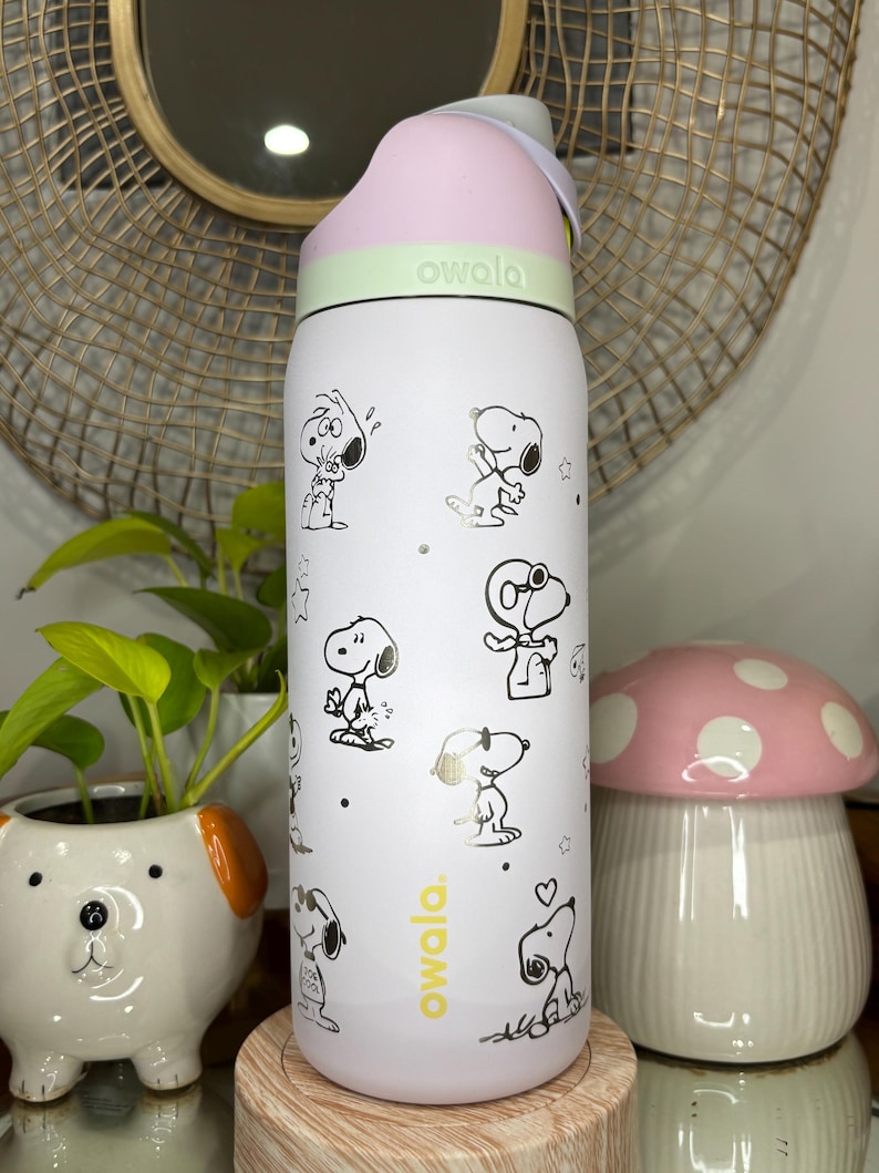 Puede incluir: Botella de agua Owala blanca con ilustraciones de Snoopy en negro y dorado. La botella tiene una tapa rosa y una banda verde claro. El logotipo de Owala est&aacute; impreso en amarillo. Una seta decorativa y una maceta con forma de perro est&aacute;n en el fondo.