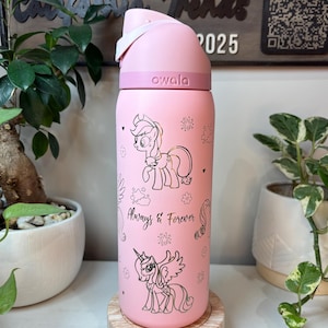 Puede incluir: Botella de agua Owala rosa con tapa abatible blanca. La botella presenta ilustraciones doradas de ponis de dibujos animados y el texto "Always & Forever". La botella es cil&iacute;ndrica y se apoya en un posavasos de madera.