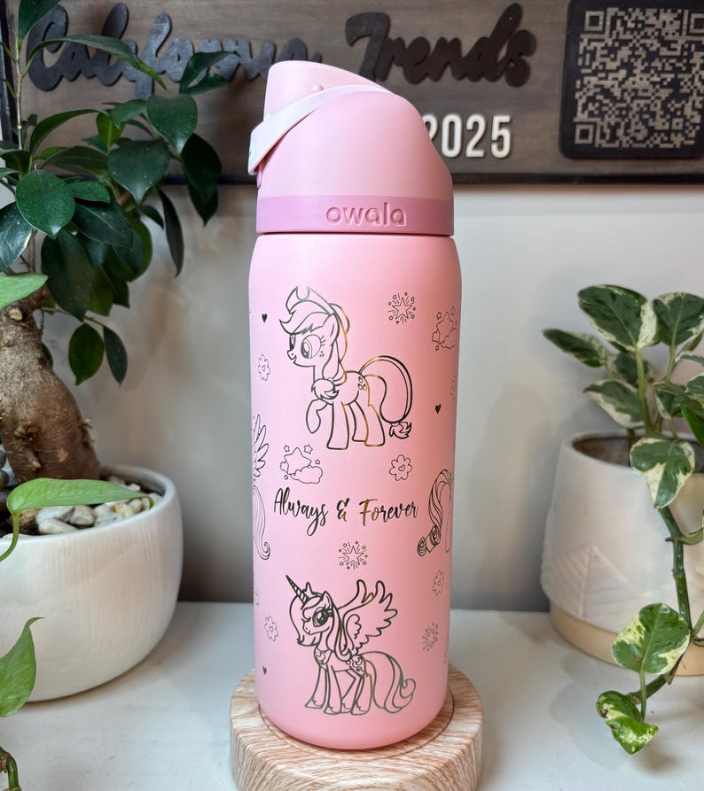 Puede incluir: Botella de agua Owala rosa con tapa abatible blanca. La botella presenta ilustraciones doradas de ponis de dibujos animados y el texto "Always & Forever". La botella es cil&iacute;ndrica y se apoya en un posavasos de madera.