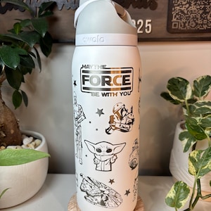 May include: White insulated water bottle with a gray lid and handle. The bottle features black and white illustrations of Star Wars characters and the phrase "MAY THE FORCE BE WITH YOU". The bottle is sitting on a wooden coaster.
