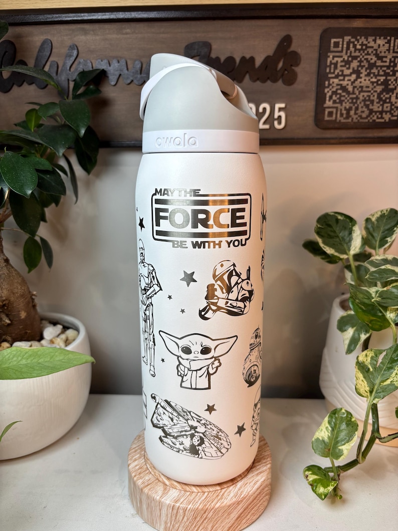 May include: White insulated water bottle with a gray lid and handle. The bottle features black and white illustrations of Star Wars characters and the phrase "MAY THE FORCE BE WITH YOU". The bottle is sitting on a wooden coaster.