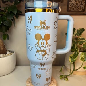 Puede incluir: Un vaso Stanley azul claro con asa, con un dise&ntilde;o dorado de Mickey Mouse y detalles de lazo. El vaso tiene una tapa transparente y ribetes dorados. El asa y la tapa son de color azul claro. El vaso est&aacute; sobre un posavasos de madera.
