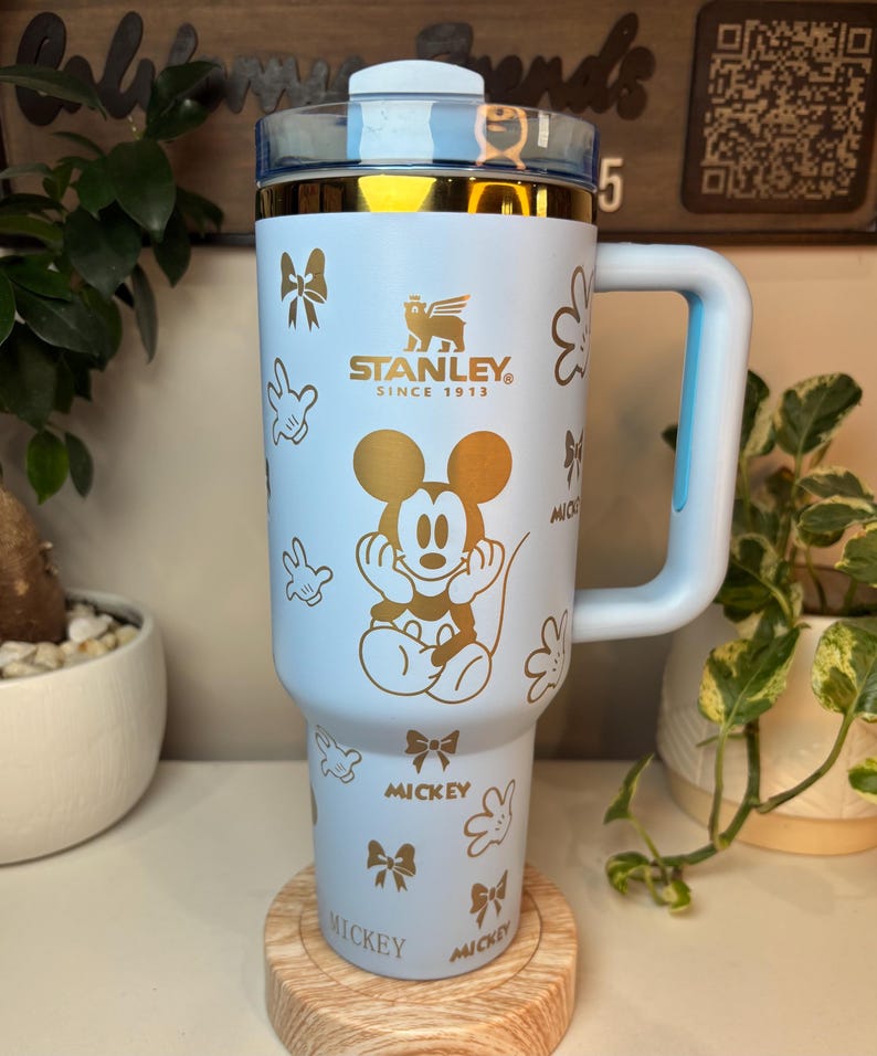 Puede incluir: Un vaso Stanley azul claro con asa, con un dise&ntilde;o dorado de Mickey Mouse y detalles de lazo. El vaso tiene una tapa transparente y ribetes dorados. El asa y la tapa son de color azul claro. El vaso est&aacute; sobre un posavasos de madera.