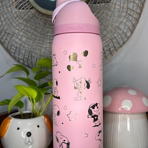 Puede incluir: Botella de agua rosa Owala con dise&ntilde;os de Snoopy, corazones y estrellas doradas. La botella tiene una tapa rosa y un asa de transporte. La palabra "owala" est&aacute; impresa en la botella. La botella est&aacute; sobre un soporte de madera.