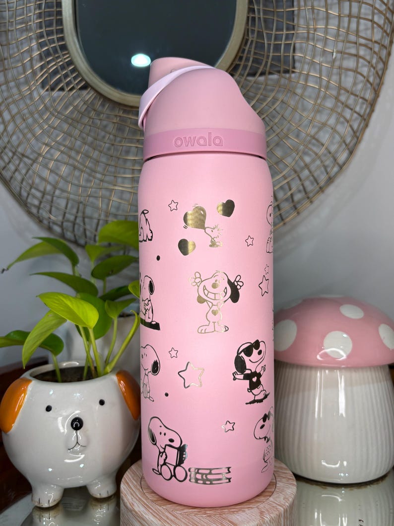 Puede incluir: Botella de agua rosa Owala con dise&ntilde;os de Snoopy, corazones y estrellas doradas. La botella tiene una tapa rosa y un asa de transporte. La palabra "owala" est&aacute; impresa en la botella. La botella est&aacute; sobre un soporte de madera.
