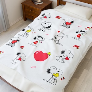 Puede incluir: Manta blanca con motivos de Snoopy y Woodstock con corazones y flores. La manta tiene un patrón repetido de Snoopy en varias poses, sosteniendo corazones y flores. También son visibles las palabras "Missing You".