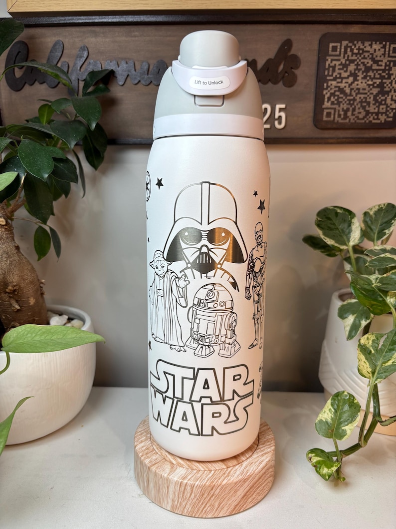 May include: White Star Wars water bottle with black character illustrations, including Darth Vader, Yoda, and R2-D2. The bottle has a gray lid with a 'Lift to Unlock' feature. The Star Wars logo is prominently displayed.