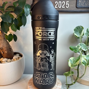 May include: A matte black Owala water bottle featuring silver laser-engraved Star Wars graphics. The design includes the text "MAY THE FORCE BE WITH YOU" and "STAR WARS," along with illustrations of Darth Vader, Yoda, R2-D2, and C-3PO.