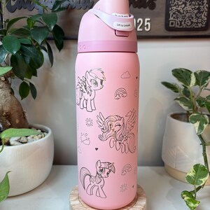 Puede incluir: Botella de agua rosa con tapa abatible, con ilustraciones de ponis de dibujos animados. La botella tiene un cuerpo rosa claro con dibujos lineales negros de ponis, nubes, arco&iacute;ris y copos de nieve. La tapa tiene el texto "Lift to Unlock".