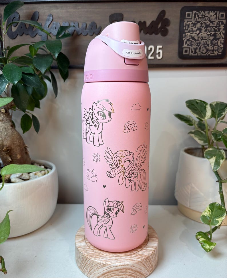 Puede incluir: Botella de agua rosa con tapa abatible, con ilustraciones de ponis de dibujos animados. La botella tiene un cuerpo rosa claro con dibujos lineales negros de ponis, nubes, arco&iacute;ris y copos de nieve. La tapa tiene el texto "Lift to Unlock".