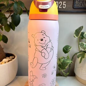 Op de afbeelding: Een lichtroze waterfles met een lijntekening van Winnie de Poeh, Knorretje, bijen en bloemen. De fles heeft een geel en oranje deksel met een vergrendelingsmechanisme met de tekst Lift to Unlock in witte letters.
