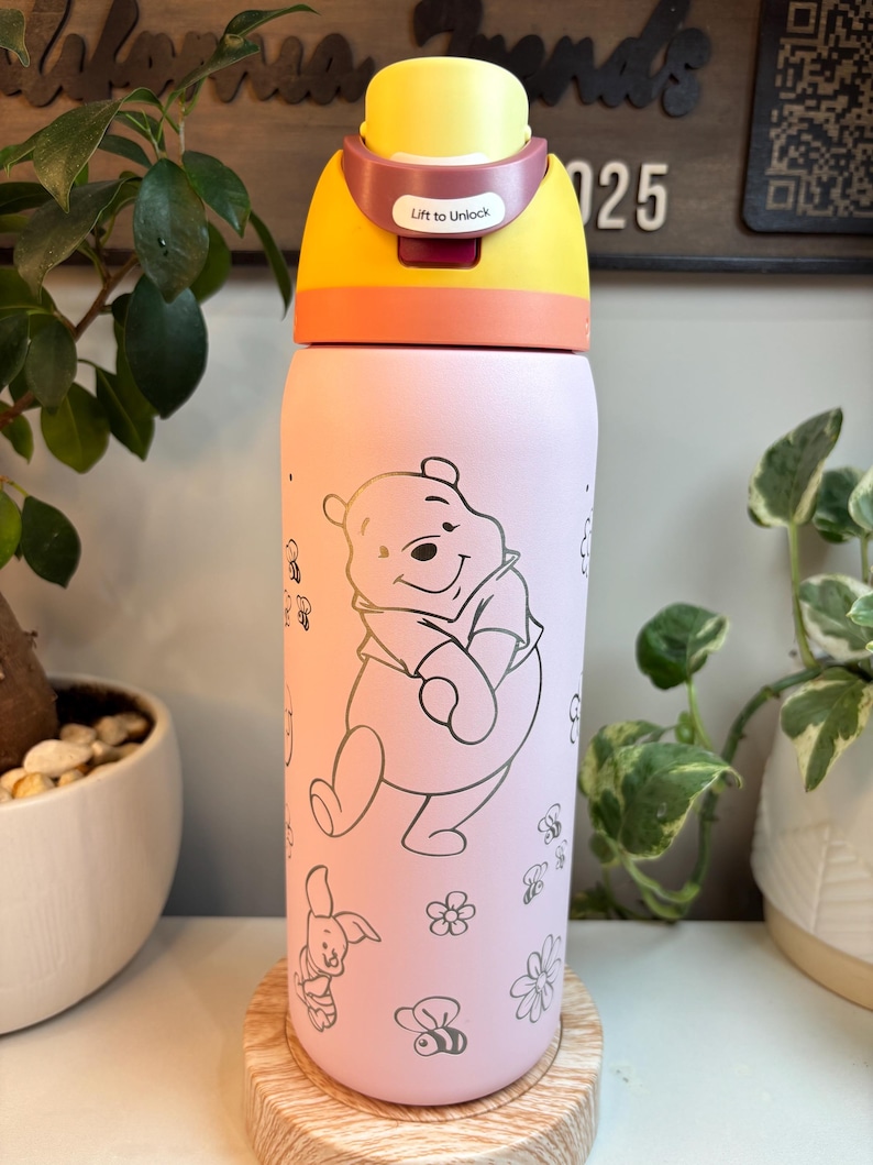 Op de afbeelding: Een lichtroze waterfles met een lijntekening van Winnie de Poeh, Knorretje, bijen en bloemen. De fles heeft een geel en oranje deksel met een vergrendelingsmechanisme met de tekst Lift to Unlock in witte letters.