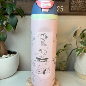 May include: A pink and blue water bottle featuring Snoopy illustrations. The bottle has a light pink body with Snoopy graphics, a blue top, and a pink locking mechanism that reads "Lift to Unlock". The bottle is on a wooden coaster.