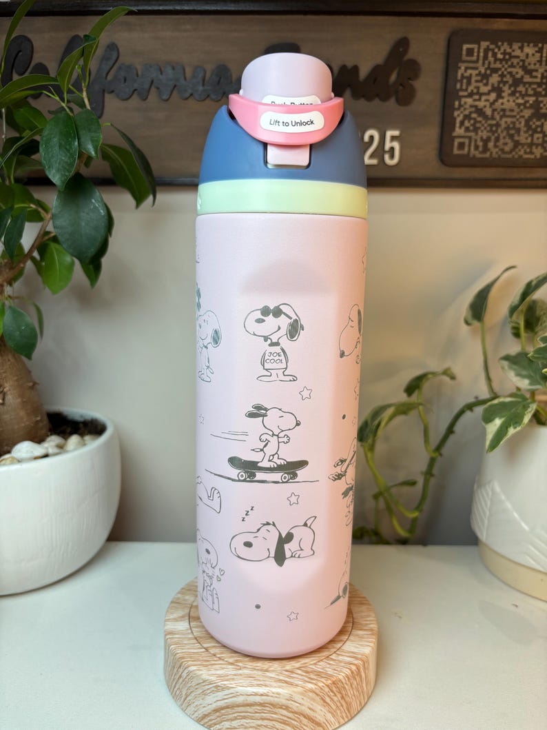 May include: A pink and blue water bottle featuring Snoopy illustrations. The bottle has a light pink body with Snoopy graphics, a blue top, and a pink locking mechanism that reads "Lift to Unlock". The bottle is on a wooden coaster.