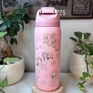 Puede incluir: Botella de agua aislada rosa con ilustraciones de ponis de dibujos animados. La botella tiene una tapa abatible y un cuerpo rosa claro. El dise&ntilde;o incluye contornos dorados de ponis, arco&iacute;ris y estrellas. La botella est&aacute; sobre un posavasos de madera.