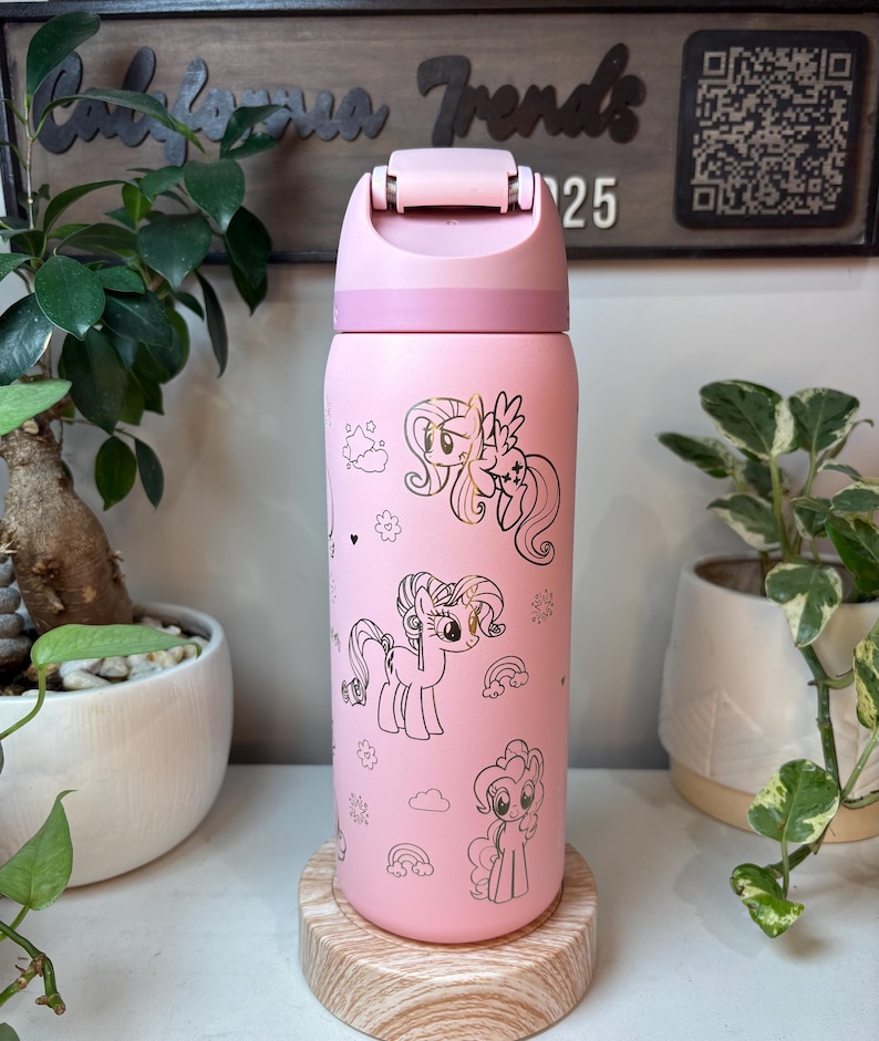Puede incluir: Botella de agua aislada rosa con ilustraciones de ponis de dibujos animados. La botella tiene una tapa abatible y un cuerpo rosa claro. El dise&ntilde;o incluye contornos dorados de ponis, arco&iacute;ris y estrellas. La botella est&aacute; sobre un posavasos de madera.